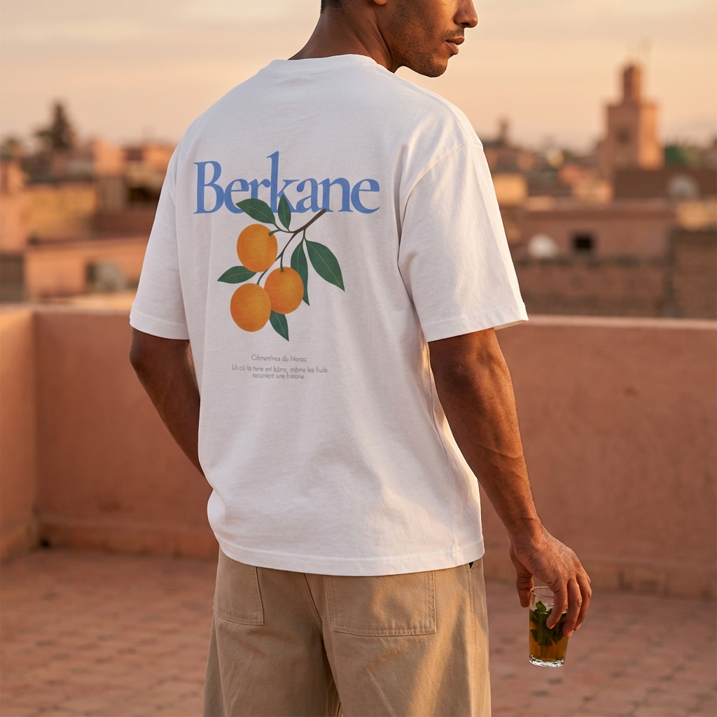 Man wearing a Berkane orange citrus inspired graphic t-shirt standing against a plain background. standing on a rooftop with a cityscape in the background.