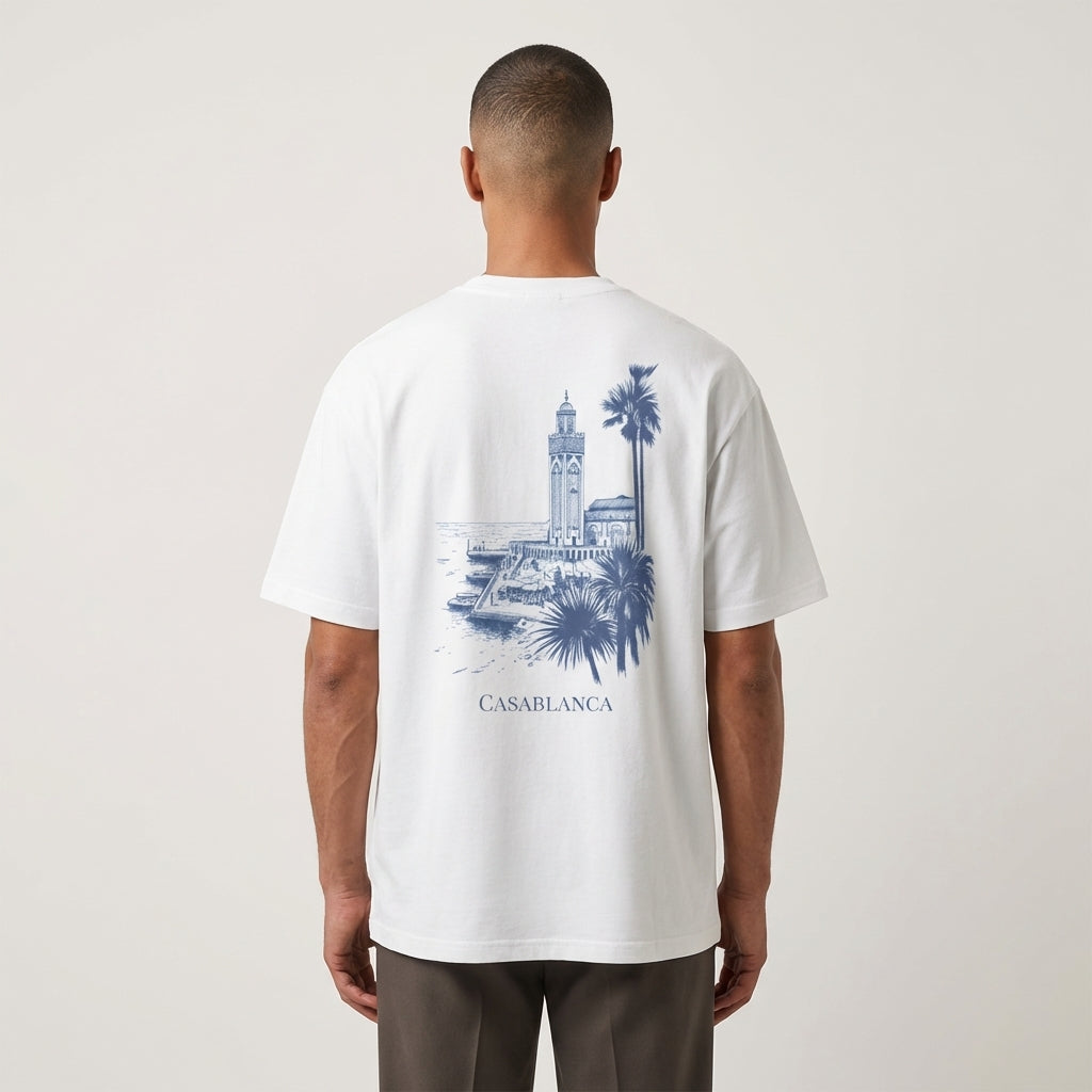 Person wearing a white t-shirt featuring a detailed blue architectural sketch of the Hassan II Mosque and palm trees, minimalist luxury streetwear fit set against a plain background