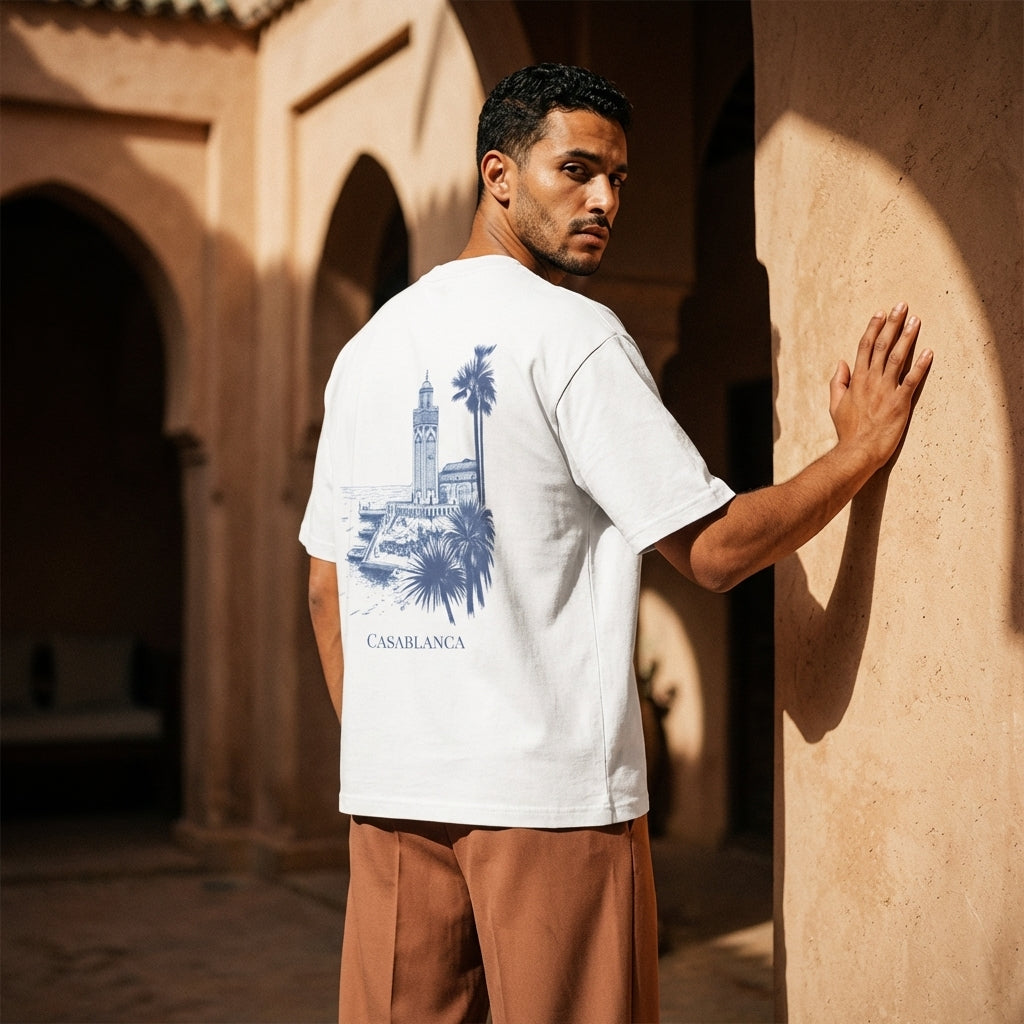 Person wearing a white t-shirt featuring a detailed blue architectural sketch of the Hassan II Mosque and palm trees, minimalist luxury streetwear fit set against a plain background, standing in a sunlit archway.