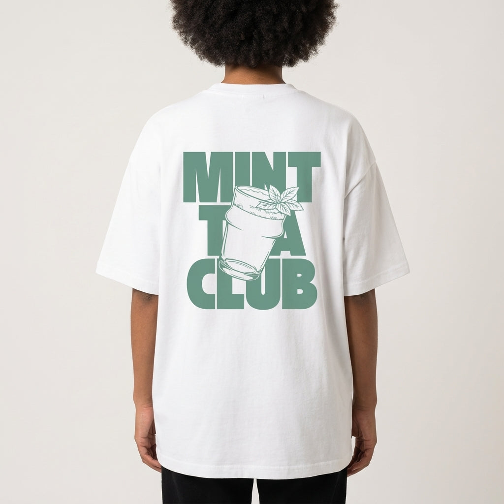Woman wearing a white heavy cotton tee with a clean sage-green graphic featuring a tea glass, fresh mint leaf, and bold 'MINT TEA CLUB' typography, minimalist Moroccan streetwear vibe against a plain background.
