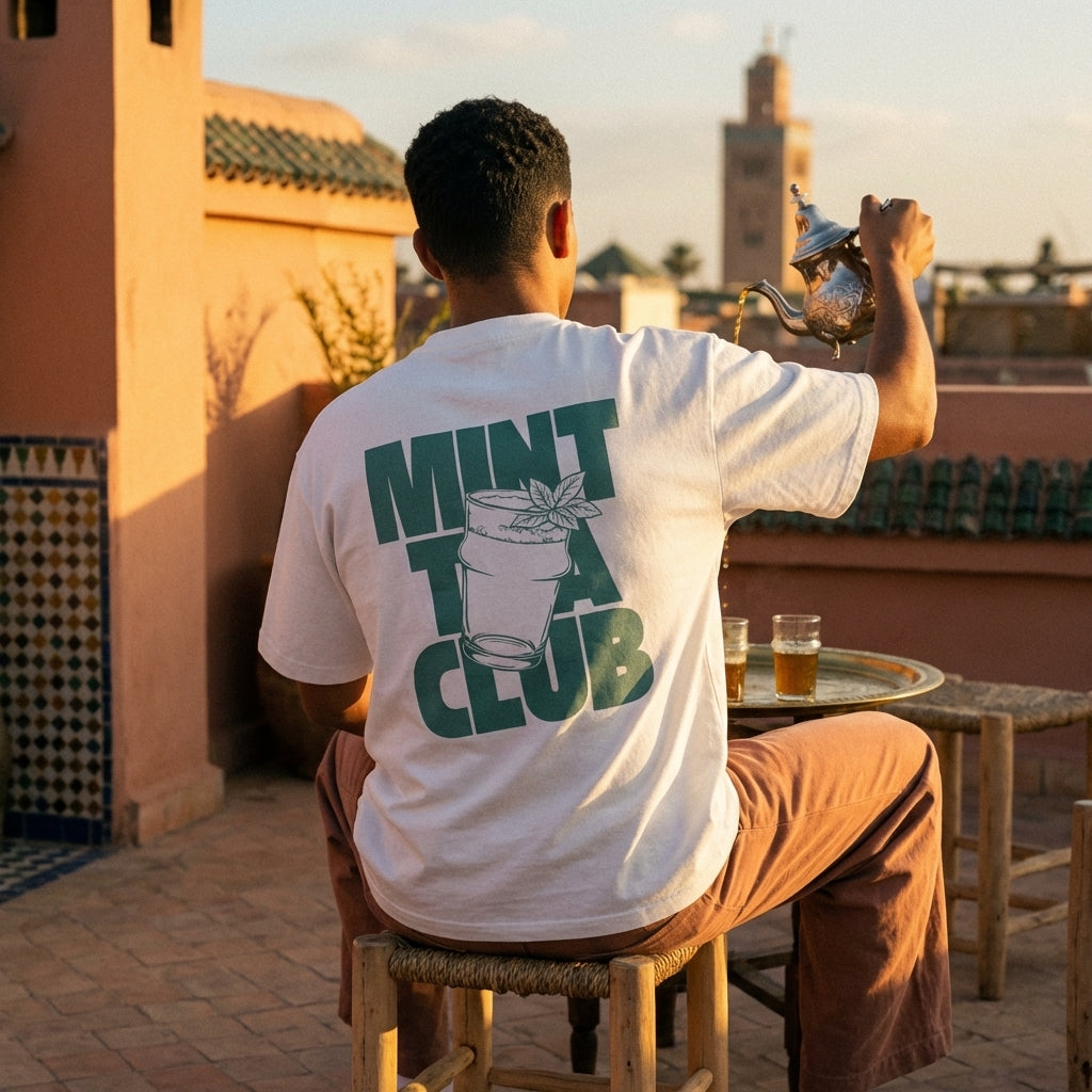 Man sitting on a rooftop with a view, wearing a white heavy cotton tee with a clean sage-green graphic featuring a tea glass, fresh mint leaf, and bold 'MINT TEA CLUB' typography, minimalist Moroccan streetwear vibe.