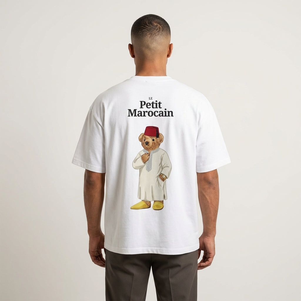 Model wearing a white t-shirt featuring a detailed illustration of a teddy bear wearing a traditional Moroccan Djellaba and Fez, 'Le Petit Marocain' text, premium cultural streetwear.standing against a plain background.