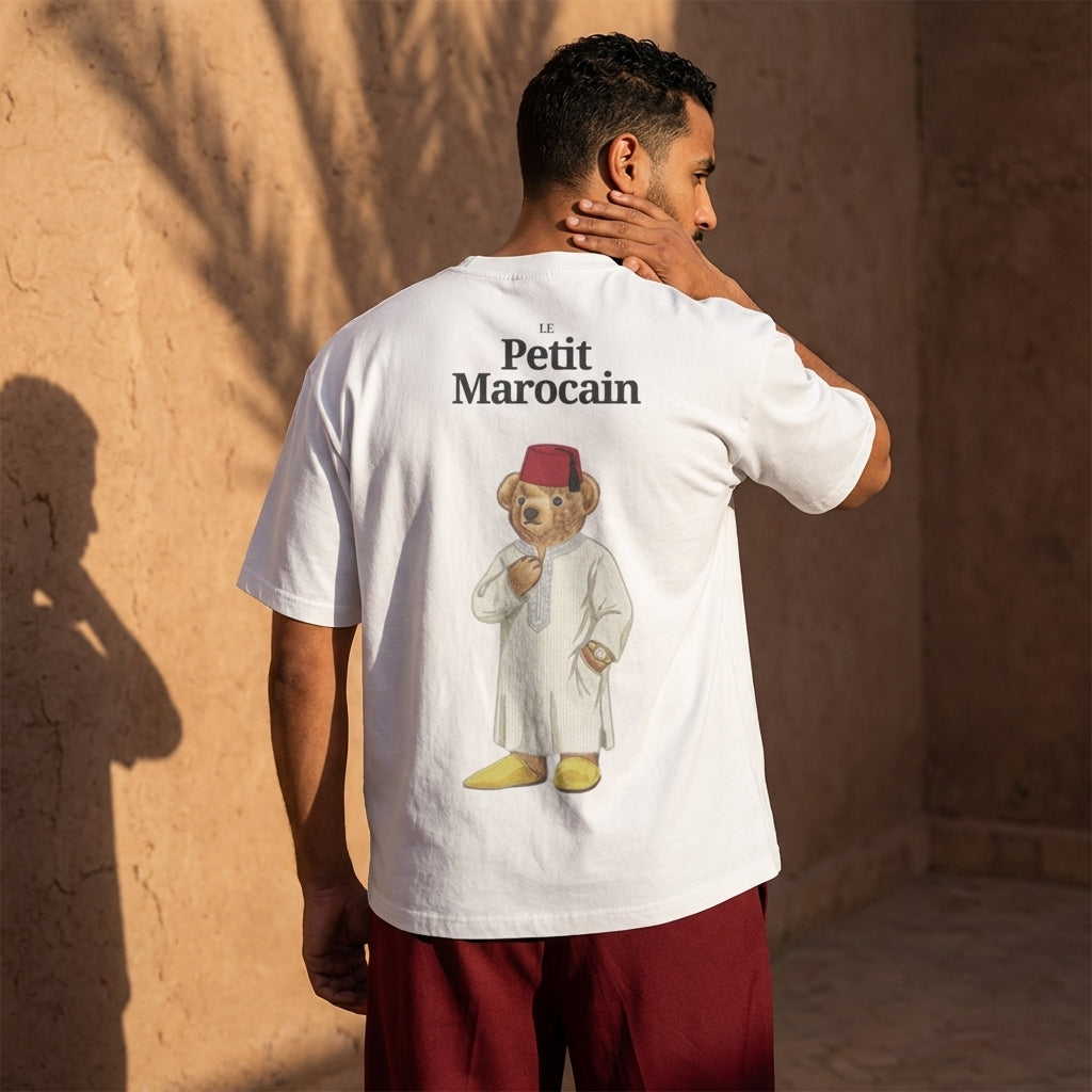 Model wearing a white t-shirt featuring a detailed illustration of a teddy bear wearing a traditional Moroccan Djellaba and Fez, 'Le Petit Marocain' text, premium cultural streetwear, standing against a warm-toned wall.