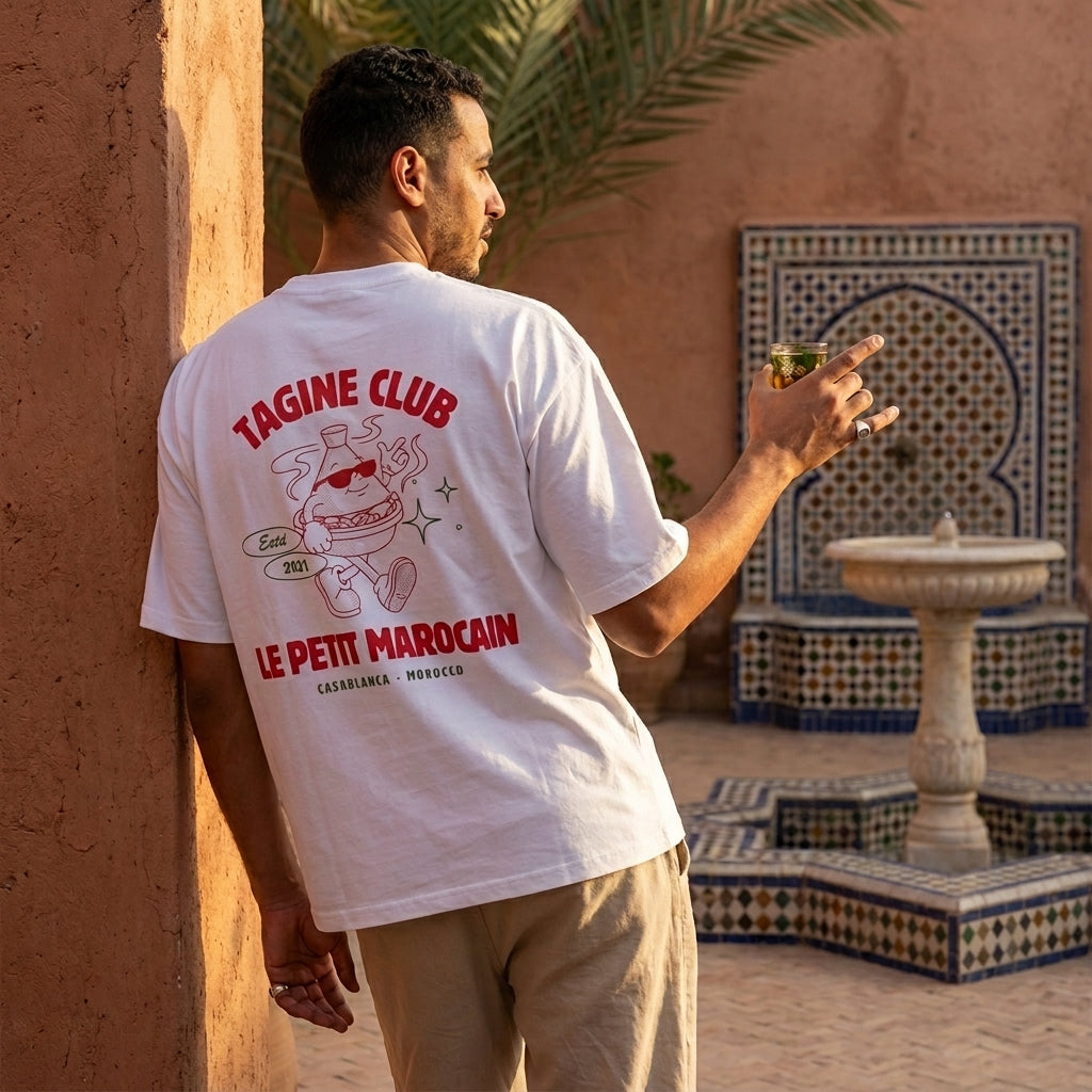 Man wearing a heavyweight t-shirt featuring a retro cartoon mascot of a walking tagine with sunglasses, 'TAGINE CLUB' red text, and 'LE PETIT MAROCAIN' branding, vintage streetwear fit and standing in a decorative outdoor setting.